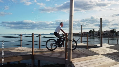 Senior man riding a mountain bike along a beach walkway on a sunny day with some clouds. Lateral camera tracking