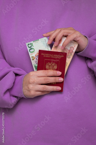 Female hands hold red Russian biometric passport, paper cash ruble and Georgian lari. Paper bill. 5000 rubles. 50 GEL. Woman in purple lilac sweatshirt with long sleeve. Immigration, travel, tourism