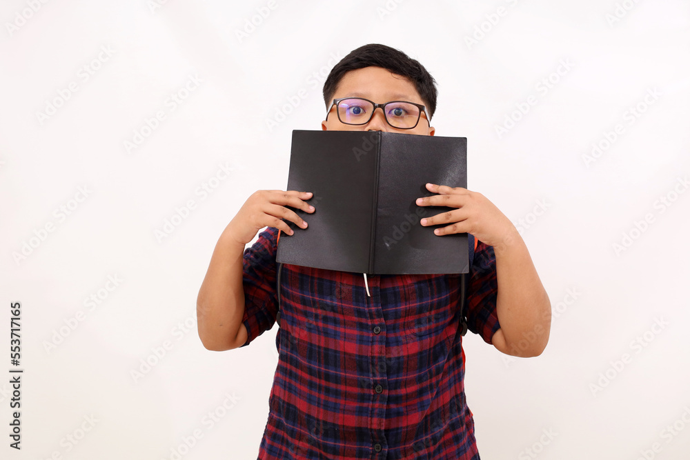 Shocked asian school boy standing while covering part of the face with his book. Isolated on white background