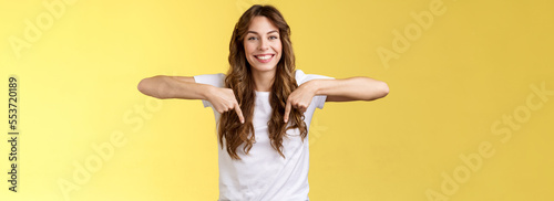 Friendly cheerful excited happy smiling girl long natural brunette haircut grinning joyfully pointing down index fingers show friend great choice introduce promo advice click link yellow background