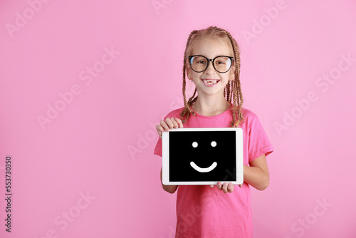 Smiling adorable little girl holds tablet on pink background. Caucasian school kid in glasses showing screen with happy emoji and looking in camera. Advertisement blank. Pupil promote product