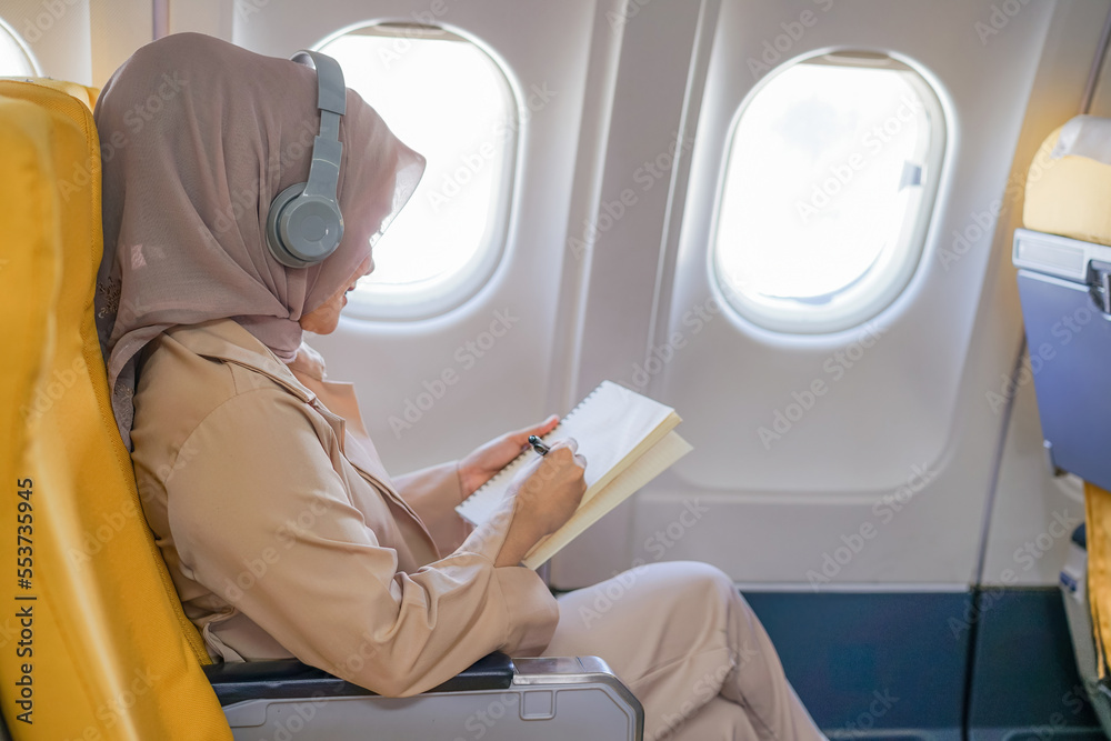 Beautiful asian muslim woman passengers wearing wireless headphone and writing on notebook. Beautiful asian muslim woman passengers wearing wireless headphone and writing on notebook.