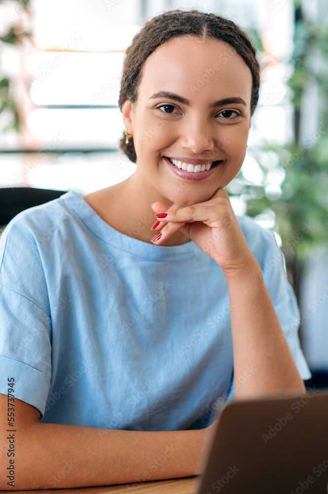 Vertical portrait of beautiful successful confident mixed race young woman, office employee, creative manager, recruitment, real estate agent, sit at workplace in modern office, looks at camera, smile