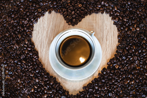 A cup of hot aromatic coffee in a saucer  against the background of scattered coffee beans in the form of a heart  on a wooden light brown table.