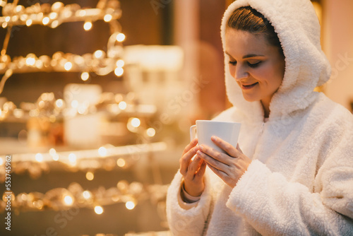 Young woman wearing white sweater drinking hot chocolate near window adorned with golden lights.