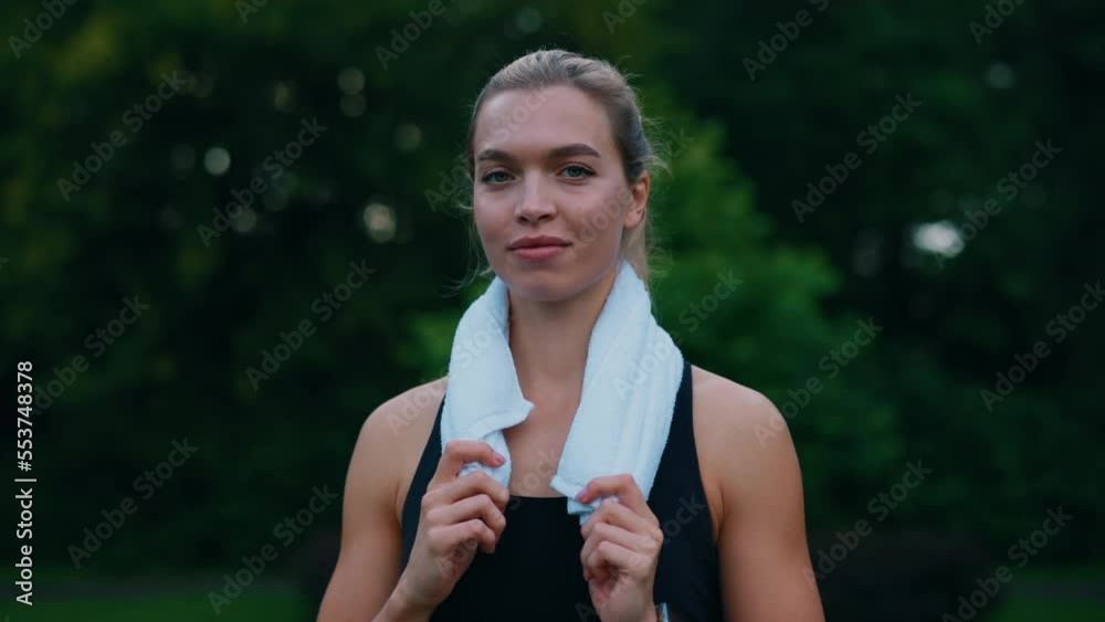 Portrait of the female runner standing lin the park before running. Caucasian athlete looking at the camera , wearing earphones carrying a towel on her neck. People and sport concept.