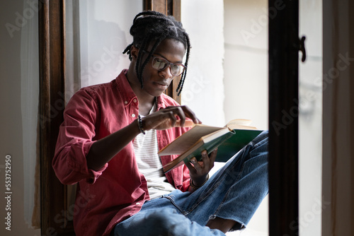 Focused emotionless African American man in casual wear spending free time reading favorite author book. Attentive student preparing for exam studying textbooks from school library sits on windowsill photo