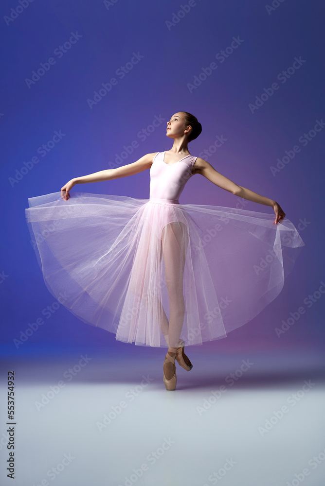 Fototapeta premium Ballet dancer. Young and incredibly beautiful ballerina is posing in point shoes and dancing. Isolated in lilac neon background. Beauty of a classical ballet art.
