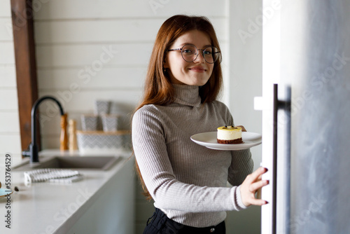 A hungry young woman takes out a sweet cake from a home refrigerator in the kitchen, smiles positively.
