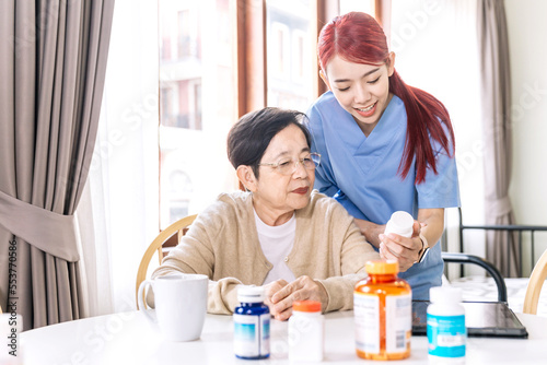 Asian woman nurse holding a medicine bottle and telling information to Asian senior woman before administering medication. Caregiver visit at home. Home health care and nursing home concept.