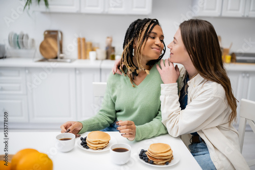 cheerful lesbian woman hugging african american girlfriend during breakfast.