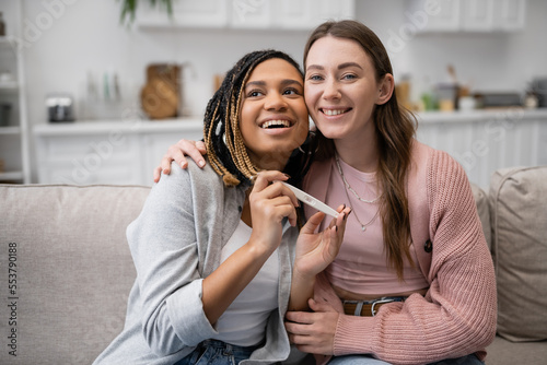 happy african american and lesbian woman holding pregnancy test near cheerful girlfriend.