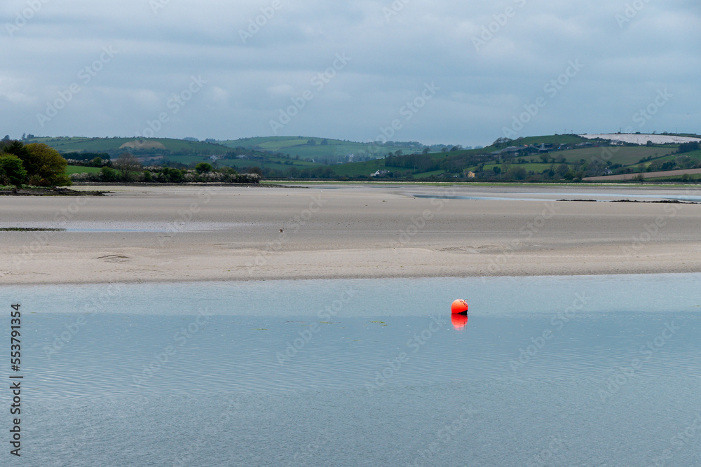 View of Clonakilty Bay at low tide. A small orange buoy is kept on a calm water surface. The sandy shore of the sea bay. The coastline on a cloudy day. View of Clonakilty Bay at low tide. A small orange buoy is kept on a calm water surface. The sandy shore of the sea bay. The coastline on a cloudy day.