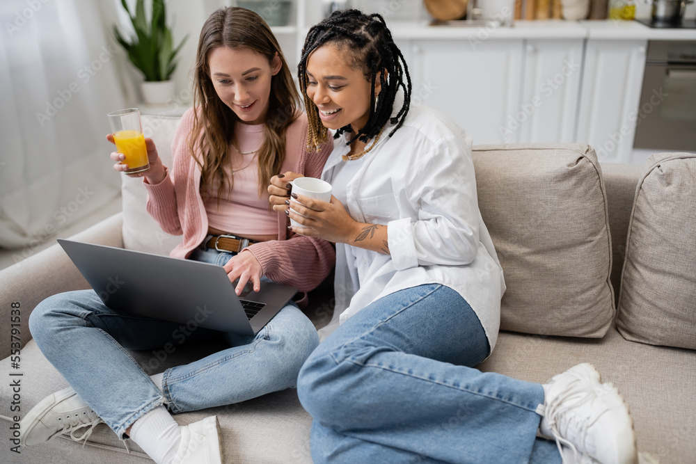 happy african american lesbian woman holding cup of coffee near girlfriend using laptop while working from home.