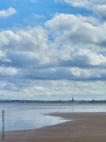 The houses, buildings and church steeple of a small coastal town form a thin ribbon dividing a huge, clouded sky from its reflection in sea and sand.