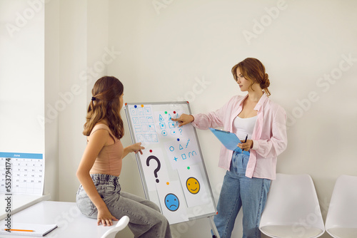 Professional female psychologist tells teenage girl more about psychology using white board. Young woman answers teenager's questions and explains while giving presentation at therapy session. photo