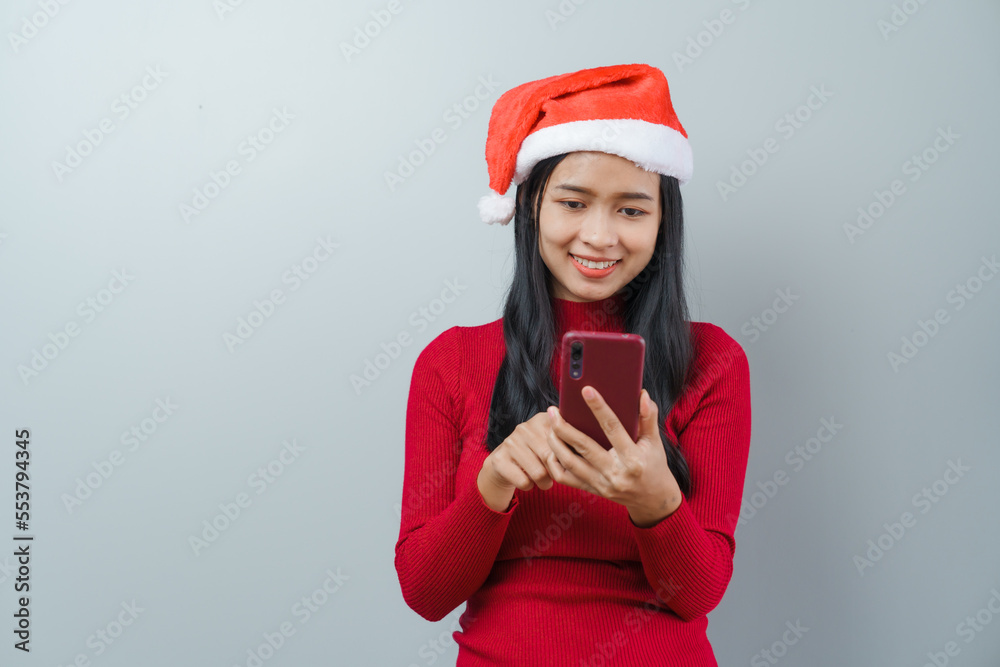 Pretty lovely lady asian thai women in red sweater and santa christmas hat isolated over grey background. Holding and using mobile smartphone or cellphone Pretty lovely lady asian thai women in red sweater and santa christmas hat isolated over grey background. Holding and using mobile smartphone or cellphone