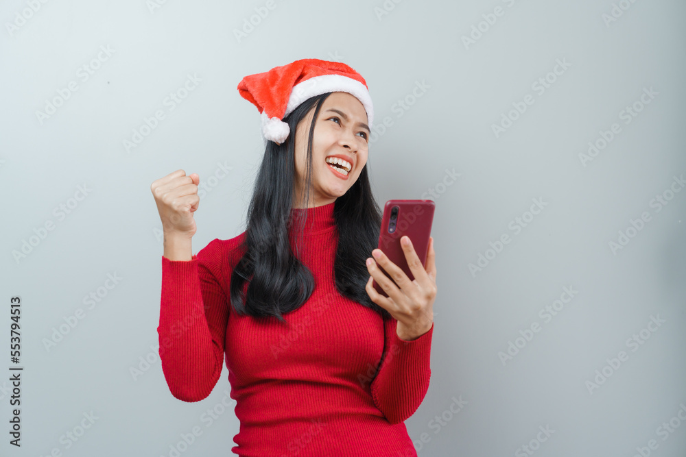 Pretty lovely lady asian thai women in red sweater and santa christmas hat isolated over grey background. Holding and using mobile smartphone or cellphone Pretty lovely lady asian thai women in red sweater and santa christmas hat isolated over grey background. Holding and using mobile smartphone or cellphone