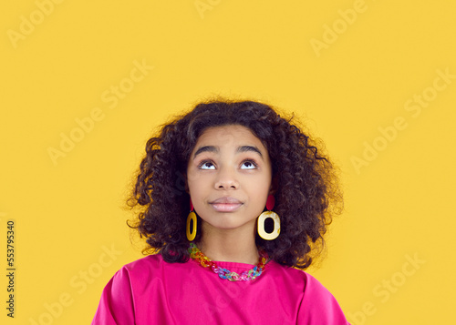 Cute african american preteen girl thinking, coming up with idea or fantasizing on yellow background. Close up of curly ethnic teen girl with colored female accessories who is looking up at copy space photo
