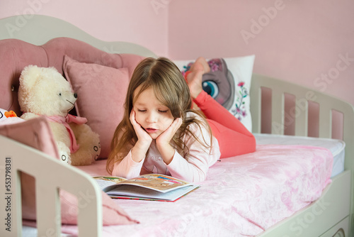 a little girl of five years with long hair lies on her children's cozy bed with toys and reads a book, looks at bright pictures photo