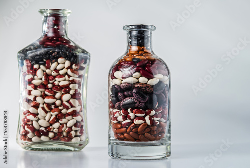 Different varieties of dry beans in glass bottles on a grey background. Selective focus.