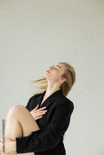 blonde model in black blazer posing with closed eyes isolated on grey.