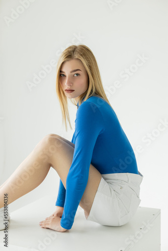 pretty and slim woman in blue turtleneck and white shorts sitting and looking at camera on grey background.