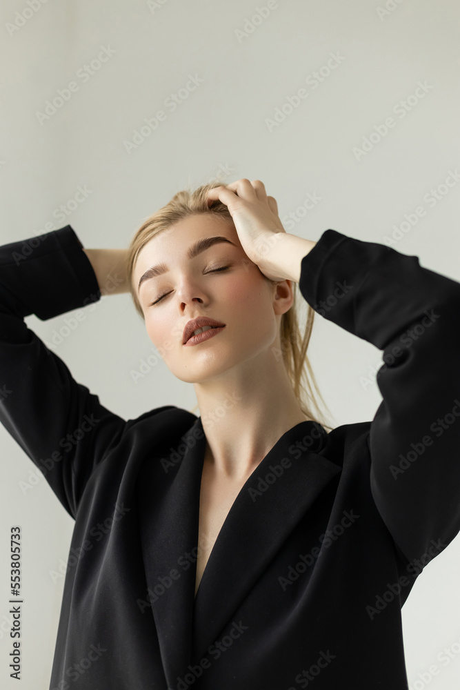 sensual young woman in black blazer touching blonde hair while posing with closed eyes isolated on grey. sensual young woman in black blazer touching blonde hair while posing with closed eyes isolated on grey.