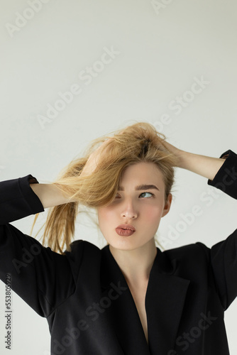 thoughtful woman in black blazer holding hair and pointing lips while looking away isolated on grey.
