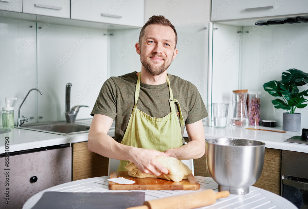 Portrait of bearded male chef baker in green apron holding dough on a cutting board with metal bowl. Making pastry or bread at home kitchen. High quality image Portrait of bearded male chef baker in green apron holding dough on a cutting board with metal bowl. Making pastry or bread at home kitchen. High quality image