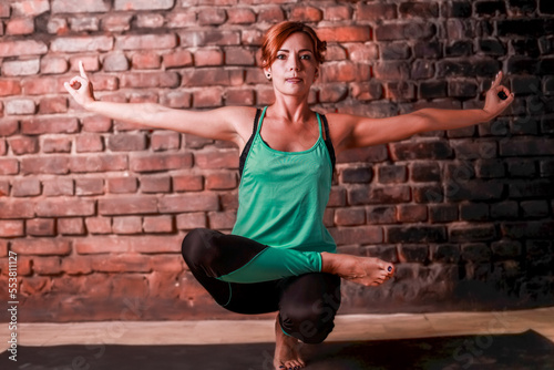 A young slender girl in black leggings and a birch T-shirt sits in an asana against the background of a brick wall.