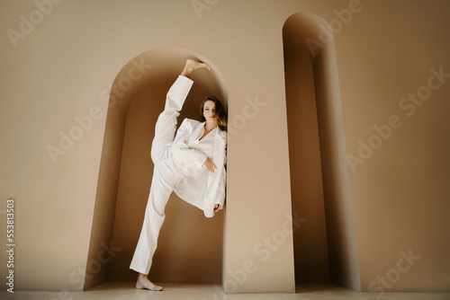 Front view of professional female model barefoot, dressed in fashionable white suit and stretching leg making twine while leaning on wall in the arch in the studio with modern interior