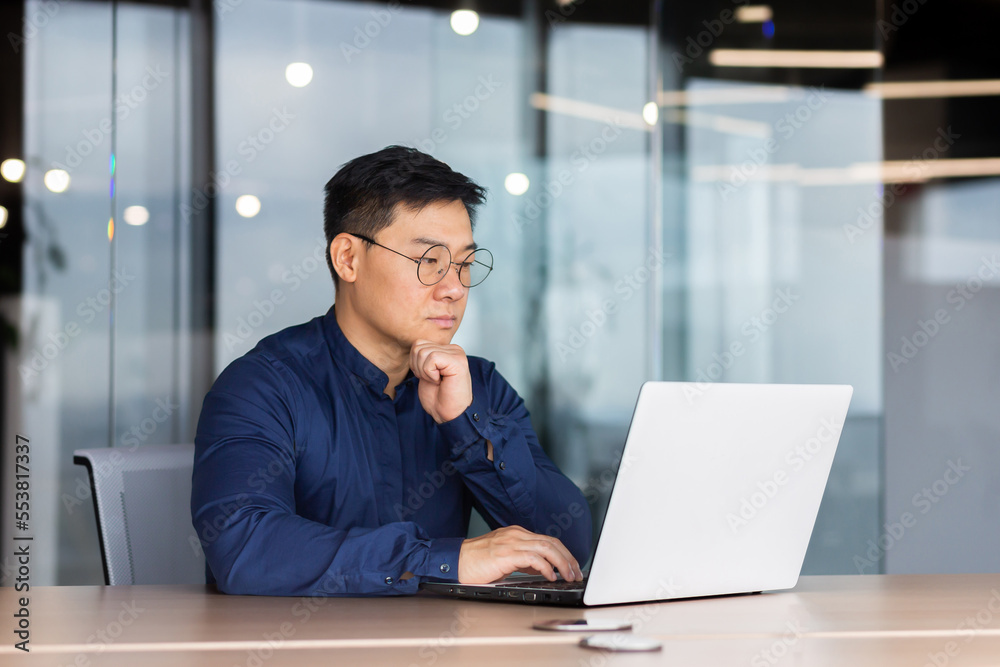 Fototapeta premium Serious and thoughtful businessman working inside office sitting at table using laptop at work, mature asian boss in shirt thinking and typing on keyboard.