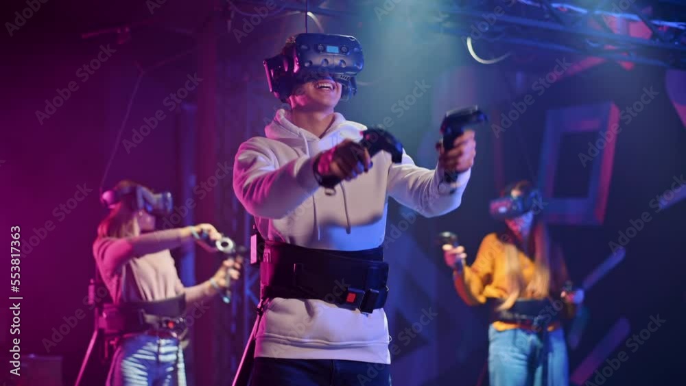 A joyful, interested boy is playing on VR treadmill with helmet, controllers and a backpack. Other people playing nearby A joyful, interested boy is playing on VR treadmill with helmet, controllers and a backpack. Other people playing nearby