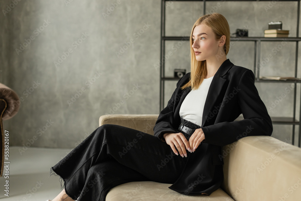 pensive blonde woman in black jeans and blazer looking away on couch in loft style living room. pensive blonde woman in black jeans and blazer looking away on couch in loft style living room.