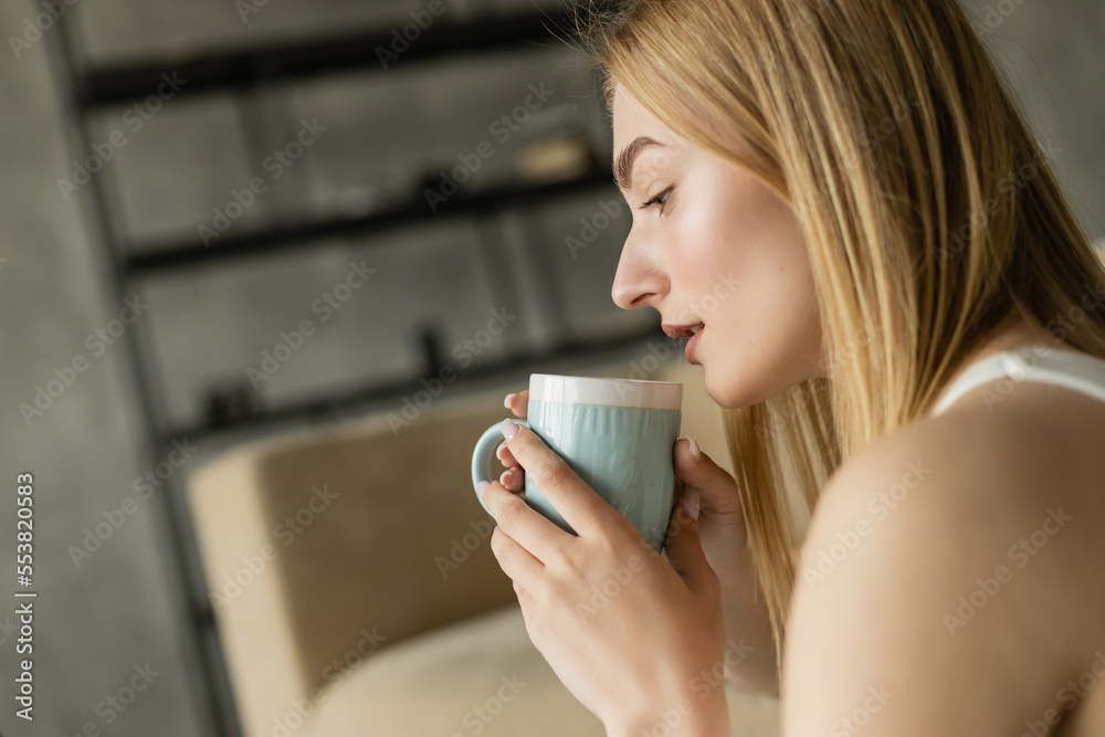 Side view of blonde woman holding cup of coffee in living room. Side view of blonde woman holding cup of coffee in living room.