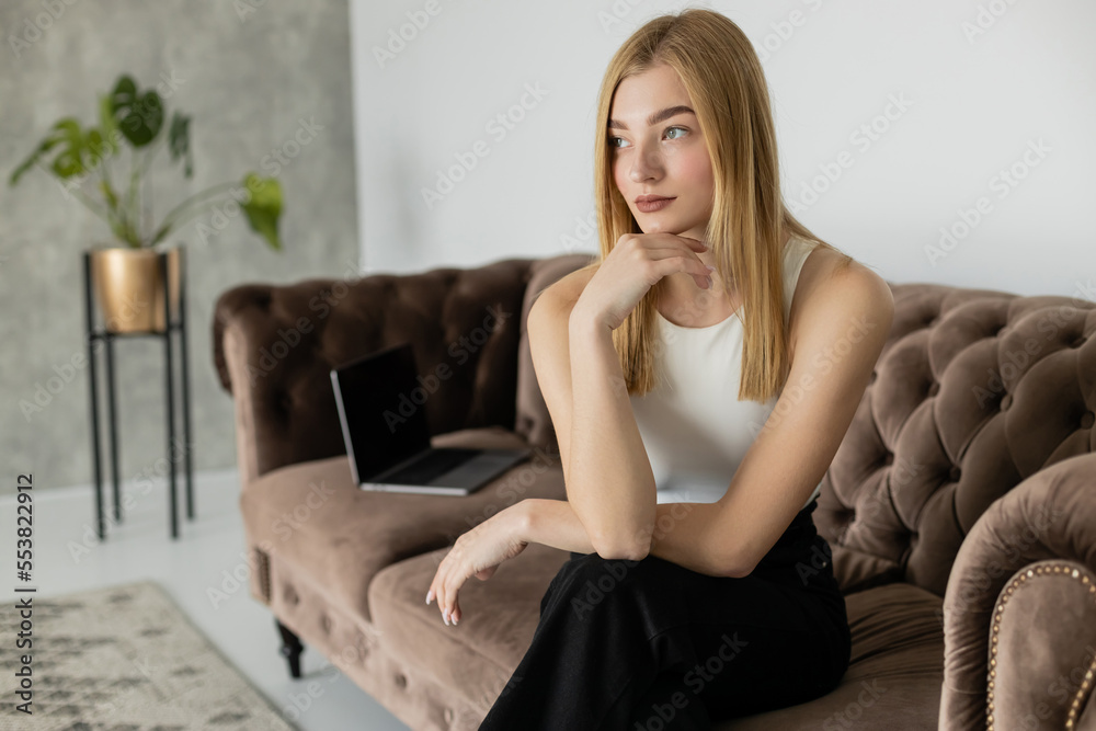 Young blonde woman in casual clothes looking away near blurred laptop on couch.