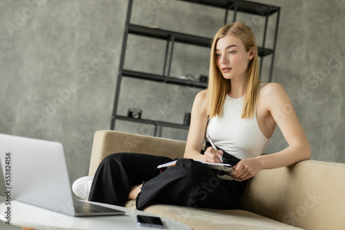 Blonde woman holding notebook near devices during online education at home.