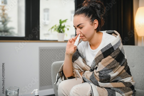 Nasal spray in hand of sick young woman sitting on couch. Allergic rhinitis symptoms and treatment