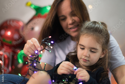 Cute little girl interacting interestedly with the Christmas lights, sitting on her mother's lap who teaches her