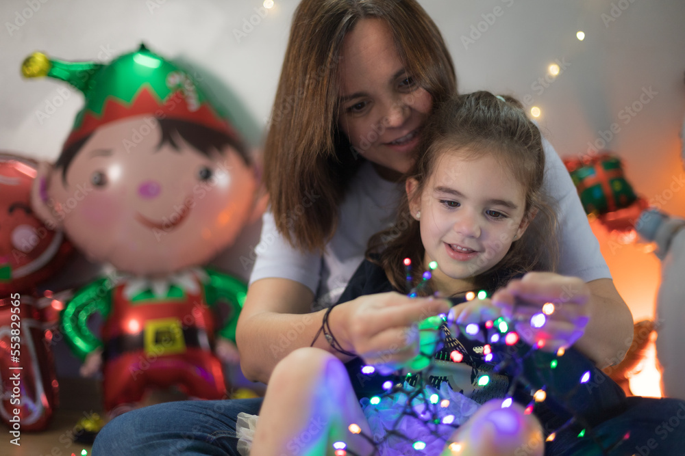 Cute little girl interacting interestedly with the Christmas lights, sitting on her mother's lap who teaches her