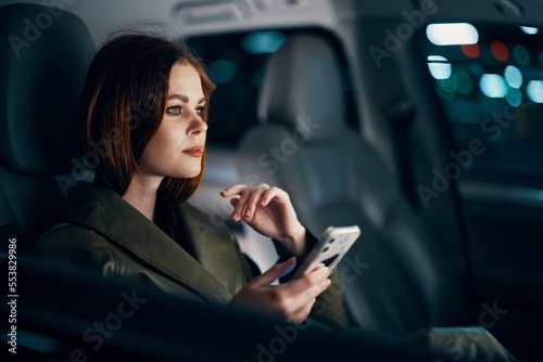  stylish, luxurious woman in a leather coat sitting in a black car at night on the passenger seat, looking thoughtfully to the side, holding her hand near her face, and the other holding a smartphone © SHOTPRIME STUDIO