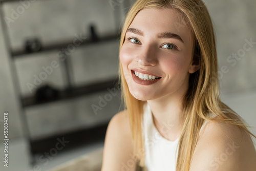 Portrait of young blonde woman smiling at camera in blurred living room. Free Download Portrait of young blonde woman smiling at camera in blurred living room. - GFXLoad