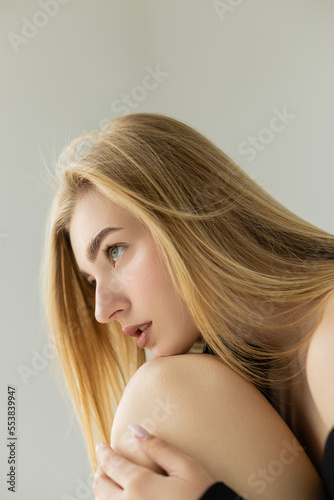 sexy blonde woman with natural makeup leaning on knee while looking away isolated on grey.