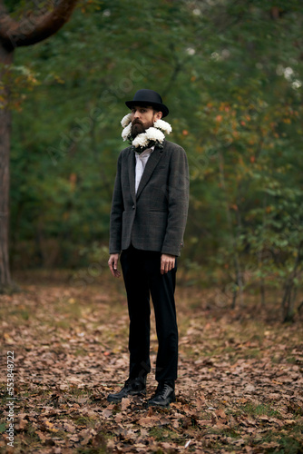 The surreal theme - a bearded man with a twisted moustache, in a blazer and hat with a bouquet of white flowers around his neck, in forest. © Vladibulgakov