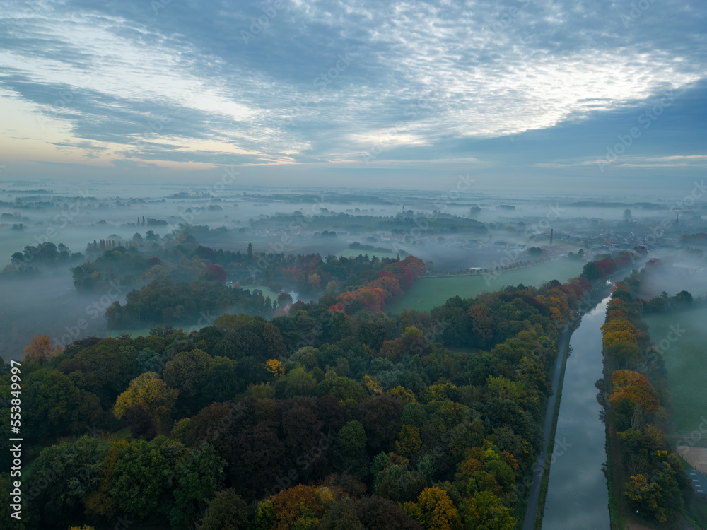 Aerial view of foggy trees in field at colorful sunrise in autumn. Colorful landscape with forest in low clouds, river, meadow in fog, orange sky with sun in the morning in fall. Top view. Nature