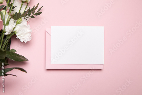 Holiday greeting card mockup with envelope and white flowers on pink background, top view, flat lay. Blank wedding invitation card mockup and floral decor