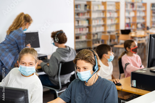 Group of young girls and boys in face masks sitting in computer classroom of library and exercising.