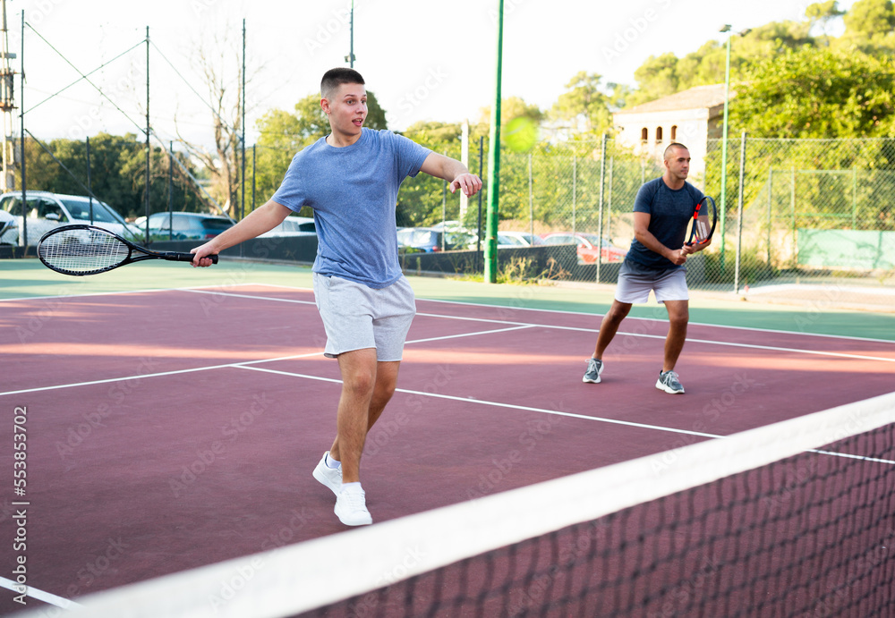 Obraz premium Young teenage tennis player training on court with male partner. Boy using racket to hit ball.
