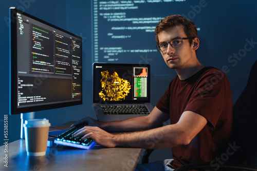 Portrait of Intelligent male programmer working on pc writing brand new code at his home office, looking at camera on background with digital wall with application info page. Data science concept